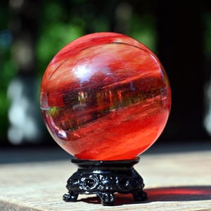 Peut inclure: Une grande boule de cristal rouge avec un motif tourbillonnant, posée sur un support noir orné. La sphère est translucide, laissant passer la lumière. Le support a un design floral décoratif.