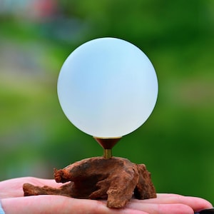 Könnte beinhalten: Eine dekorative Lampe mit einer mattweißen Glaskugel auf einem Messingstiel, montiert auf einem Stück strukturiertem Holz. Das Design der Lampe ist minimalistisch und organisch, mit Fokus auf natürlichen Materialien.