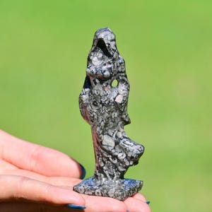以下が含まれることがあります： グレーと白の色合いの、フード付きの人物の小さな彫刻された石像。像は、ぼやけた緑の背景に対して、人の手の中に保持されています。この置物は、テクスチャ加工された表面とユニークな抽象的なデザインを持っています。