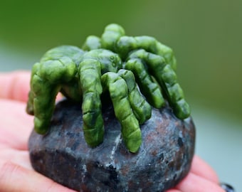May include: A detailed, green spider sculpture with eight legs, perched on a dark, textured stone. The spider is a vibrant green color, and the stone has a mottled appearance. The sculpture is held in a hand against a blurred green background.