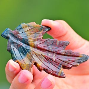 May include: A carved leaf sculpture with a detailed design. The leaf is made of a stone with a mix of colors, including blue, brown, and red. The sculpture is held in a hand against a blurred green background.