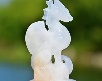 May include: A carved white dragon figurine with a detailed design. The sculpture has a stylized form with wings and a long neck. The base is a light orange color. The background is blurred with green and blue tones.