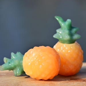 以下が含まれることがあります： 3つのパイナップルの装飾品。2つはオレンジ色で緑色の葉があり、1つはテクスチャ加工された表面の小さなオレンジ色の球体です。装飾品は、ぼやけた青色の背景の木製の表面にあります。