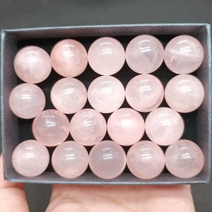 Puede incluir: Una caja de 16 esferas de cuarzo rosa. Las esferas son lisas y pulidas, y están dispuestas en un patrón de cuadrícula.