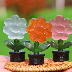 May include: Three carved flower sculptures, each with a different coloured flower: white, orange, and pink. The flowers sit atop green leaves and black pots, all resting on a woven mat. The background is a blurred green.