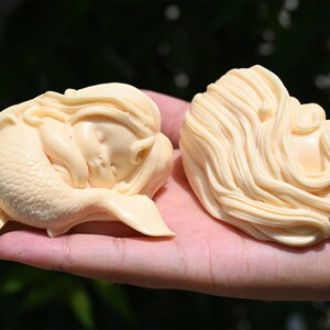 May include: Two ivory-colored mermaid figurines, one with a tail and the other with long flowing hair, are lying down with their eyes closed. The figurines are made of a smooth, polished material and have a delicate, whimsical appearance.