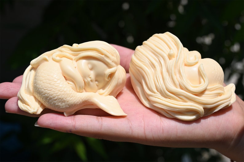 May include: Two ivory-colored mermaid figurines, one with a tail and the other with long flowing hair, are lying down with their eyes closed. The figurines are made of a smooth, polished material and have a delicate, whimsical appearance.