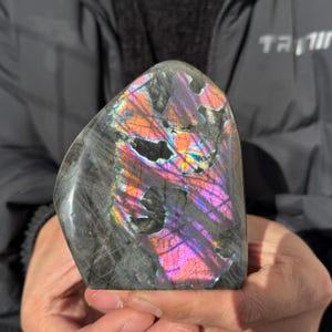 以下が含まれることがあります： 磨かれた、不規則な形のラブラドライトの石。石は、紫、ピンク、オレンジ、黄色の虹色の閃光を持つ濃い灰色のベースカラーを示しています。石は人の手で持たれています。