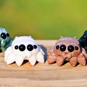 May include: Four decorative spider figurines in shades of green, white, brown, and black, each with multiple black eyes. The spiders are displayed on a wooden surface against a blurred green background.