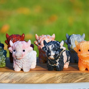May include: A collection of carved stone Highland cow figurines in various colors, including green, pink, orange, and black. The figurines are displayed on a wooden surface against a blurred green background. Each cow has detailed features.
