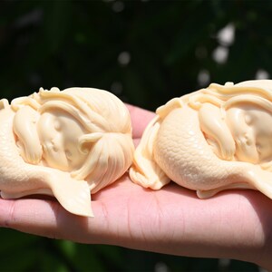 May include: Two ivory-colored mermaid figurines, each with a sleeping baby face and a curled tail, are displayed on a hand.