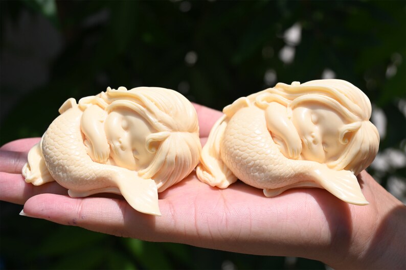 May include: Two ivory-colored mermaid figurines, each with a sleeping baby face and a curled tail, are displayed on a hand.