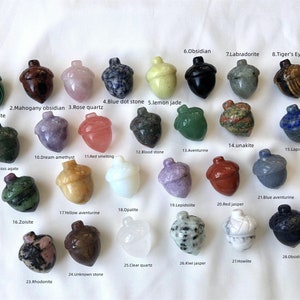 May include: Twenty-eight acorn-shaped polished stones, each carved from a different type of gemstone. The stones are arranged in rows on a white surface. The stones include malachite, mahogany obsidian, rose quartz, blue dot stone, lemon jade, obsidian, labradorite, tiger's eye, unakite, bloodstone, aventurine, lapis lazuli, dream amethyst, red smelting, moss agate, zoisite, yellow aventurine, opalite, lepidolite, red jasper, blue aventurine, fluorite, rhodonite, unknown stone, clear quartz, kiwi jasper, howlite, and obsidian.
