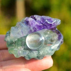 May include: A green and purple fluorite crystal carved into the shape of a clam shell with a clear crystal ball inside.