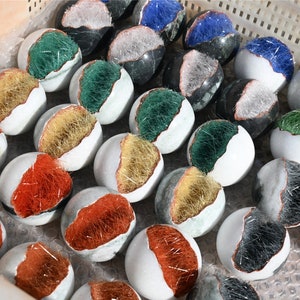 Può includere: Una collezione di pietre sferiche bianche, ciascuna divisa a metà per rivelare un interno colorato e strutturato. Gli interni variano di colore, tra cui rosso, oro, verde, blu e grigio. Le pietre sono disposte in un contenitore di plastica bianca.