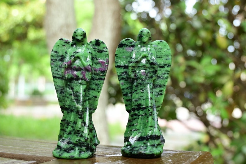 May include: Two green and black gemstone angel figurines with wings. The figurines are carved from a green stone with black and pink streaks.