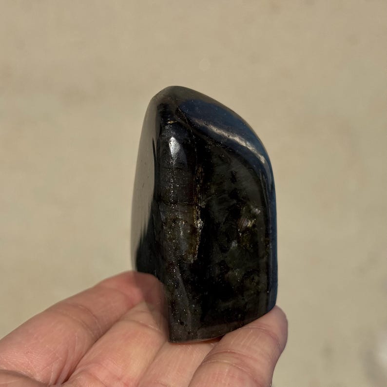 May include: A smooth, black, polished gemstone with a slightly iridescent sheen. The gemstone is shaped like a rounded rectangle and is about 2 inches tall.