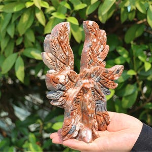May include: A carved stone eagle sculpture with outstretched wings, featuring shades of red, brown, and gray. The eagle is held in a hand against a backdrop of green foliage. The sculpture has detailed feather patterns.