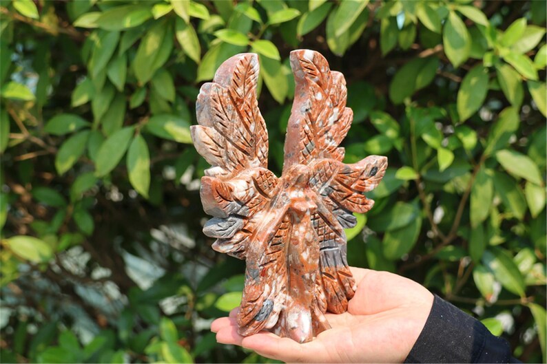 May include: A carved stone eagle sculpture with outstretched wings, featuring shades of red, brown, and gray. The eagle is held in a hand against a backdrop of green foliage. The sculpture has detailed feather patterns.