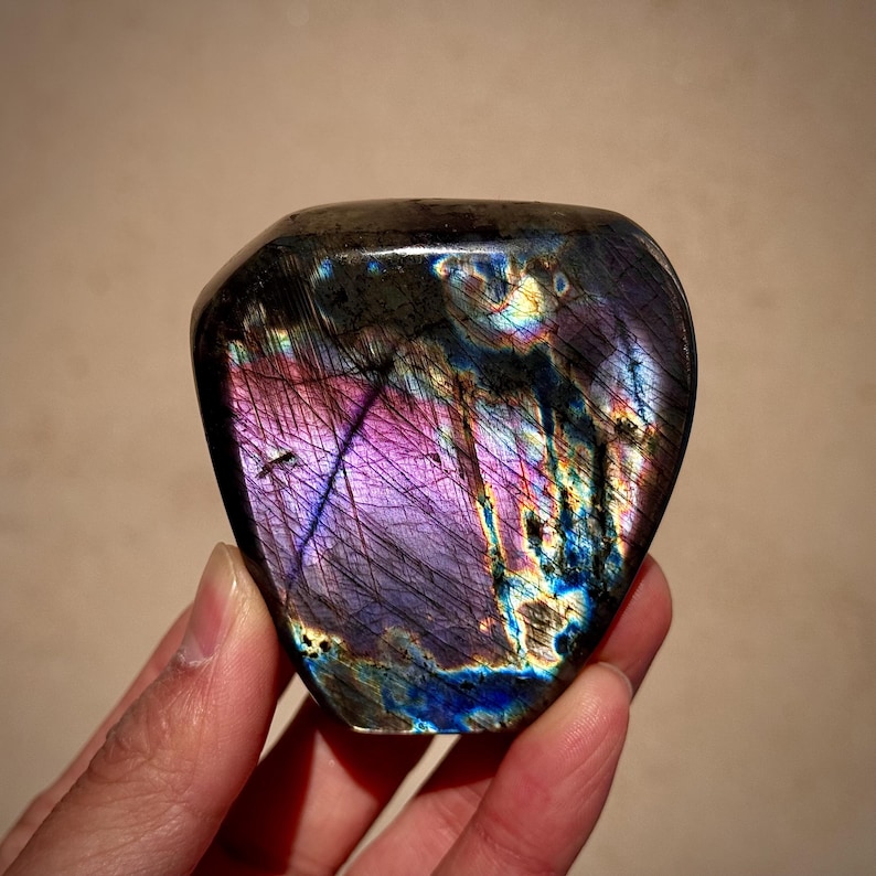 May include: A heart-shaped labradorite crystal with iridescent purple, blue, and green hues. The crystal has a smooth, polished surface and is a beautiful example of this gemstone.