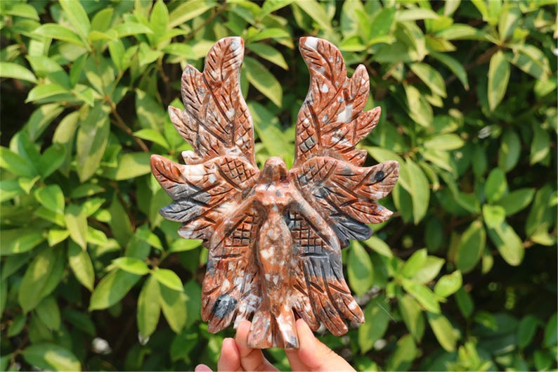 May include: A carved stone angel figurine with outstretched wings. The angel is primarily reddish-brown with gray and white accents. The wings are detailed with feather-like patterns. The background is a blur of green foliage.