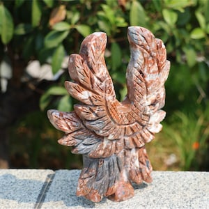 May include: A carved stone eagle sculpture with outstretched wings. The sculpture features shades of red, brown, and gray, with detailed feather carvings. The eagle stands on a small base, showcasing the craftsmanship of the artwork.