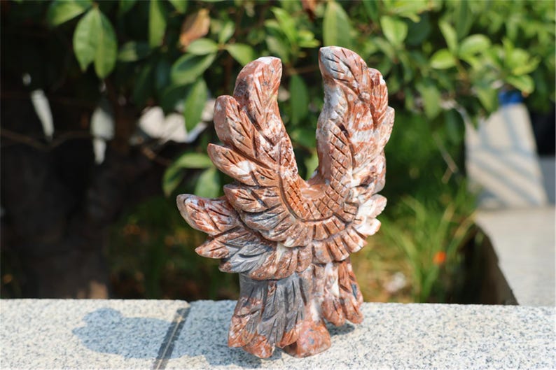 May include: A carved stone eagle sculpture with outstretched wings. The sculpture features shades of red, brown, and gray, with detailed feather carvings. The eagle stands on a small base, showcasing the craftsmanship of the artwork.