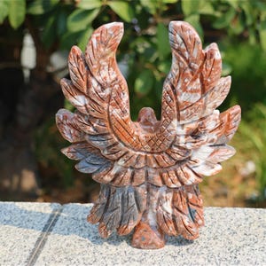 May include: A carved stone eagle sculpture with outstretched wings. The sculpture is primarily reddish-brown with white and gray veining, and is set against a blurred green background. The eagle's wings are detailed with carved feather patterns.