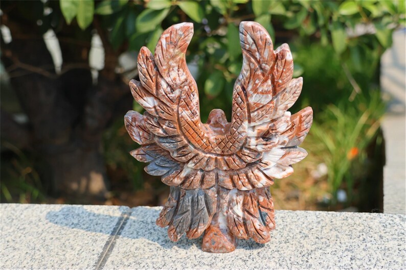 May include: A carved stone eagle sculpture with outstretched wings. The sculpture is primarily reddish-brown with white and gray veining, and is set against a blurred green background. The eagle's wings are detailed with carved feather patterns.