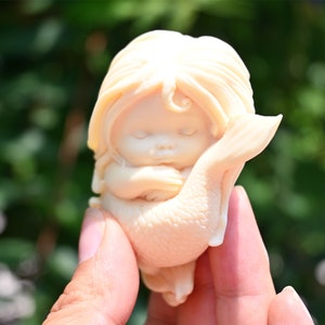 May include: A small, white ceramic figurine of a mermaid with long hair, sleeping with her arms wrapped around her tail.