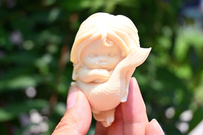 May include: A small, white ceramic figurine of a mermaid with long hair, sleeping with her arms wrapped around her tail.