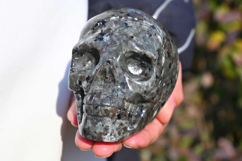 May include: A black and grey crystal skull with a detailed carving. The skull is made of a polished stone with a speckled pattern.