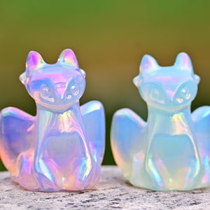 May include: Two iridescent fox ornaments with a pearlescent finish. The foxes have large ears and wings, and the colours shift from pink to blue and yellow. The ornaments are displayed on a textured surface against a blurred green background.