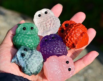 May include: A hand displays a selection of small, owl-shaped figurines in different colours. The owls are green, clear, red, purple, blue-grey, and pink, each with black eyes. The figurines have a textured surface.