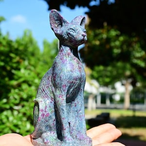 May include: A seated, stylized cat sculpture with a blue-grey and purple speckled finish. The cat has large ears and is posed in a regal, upright position. The sculpture is held in a hand against a backdrop of green foliage and a blue sky.