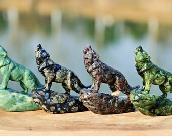 Op de afbeelding: Vier gebeeldhouwde stenen wolfsfiguren in verschillende kleuren: groen, zwart en grijs, roodbruin en nog een groen exemplaar. Elke wolf is afgebeeld terwijl hij huilt, staand op een rots. De achtergrond is wazig.