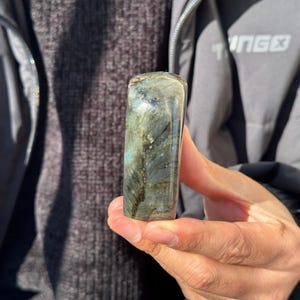 May include: A smooth, polished, dark gray and blue labradorite crystal with iridescent shimmer. The crystal is shaped like a rounded rectangle.