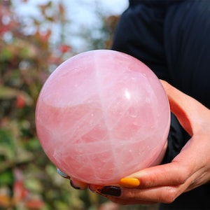 Peut inclure: Une grande sphère de quartz rose de couleur rose avec une surface lisse et polie. La sphère est tenue dans la main d'une personne.