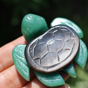 Peut inclure: Une figurine de tortue en pierre verte avec une carapace gris foncé. Le corps de la tortue est d'un vert vif et la carapace présente un motif détaillé. La figurine est tenue dans une main, avec un arrière-plan vert flou.