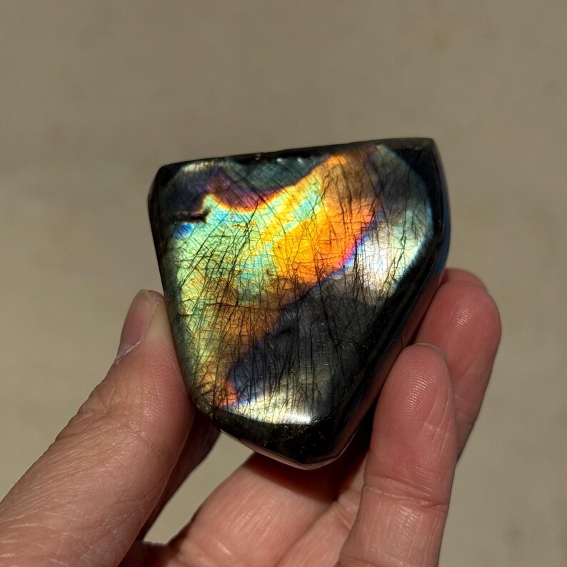 May include: A black labradorite gemstone with iridescent rainbow colors, showing a flash of orange, blue, and green. The stone is smooth and polished, with a heart-shaped form.