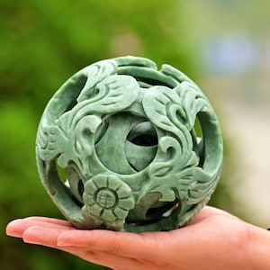 May include: A carved jade sphere with intricate floral and swirling patterns. The light green stone is sculpted with openwork designs, revealing the interior. The sphere is held in a hand against a blurred green background.