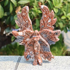 May include: A carved stone sculpture of a winged figure with outstretched arms. The sculpture features intricate details in shades of red, brown, and gray. The figure is standing on a gray surface, with green foliage in the background.