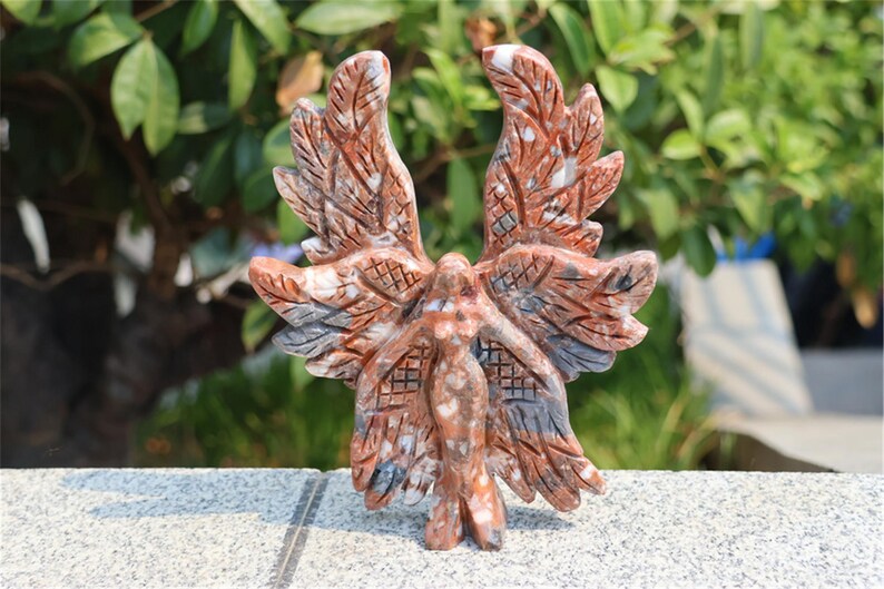 May include: A carved stone sculpture of a winged figure with outstretched arms. The sculpture features intricate details in shades of red, brown, and gray. The figure is standing on a gray surface, with green foliage in the background.