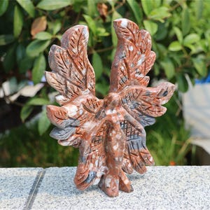 May include: A carved stone sculpture of an eagle with outstretched wings. The sculpture is primarily reddish-brown with white and gray accents. The eagle is standing on a gray surface, with a blurred green background of foliage.