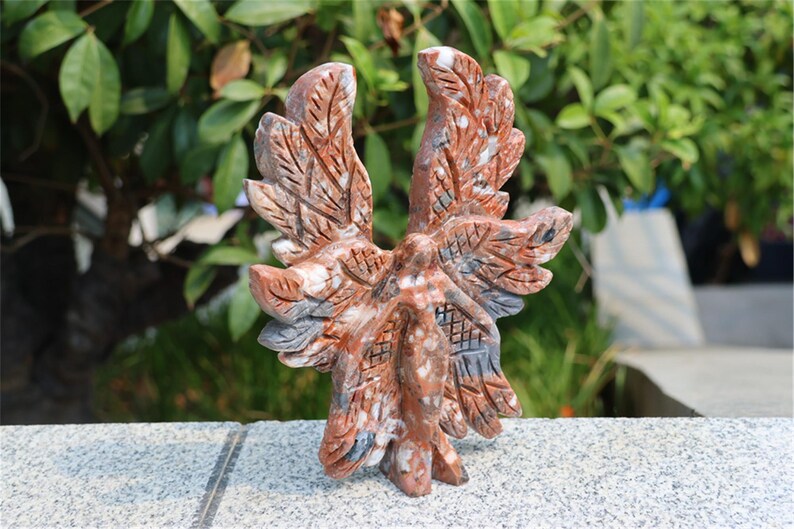May include: A carved stone sculpture of an eagle with outstretched wings. The sculpture is primarily reddish-brown with white and gray accents. The eagle is standing on a gray surface, with a blurred green background of foliage.