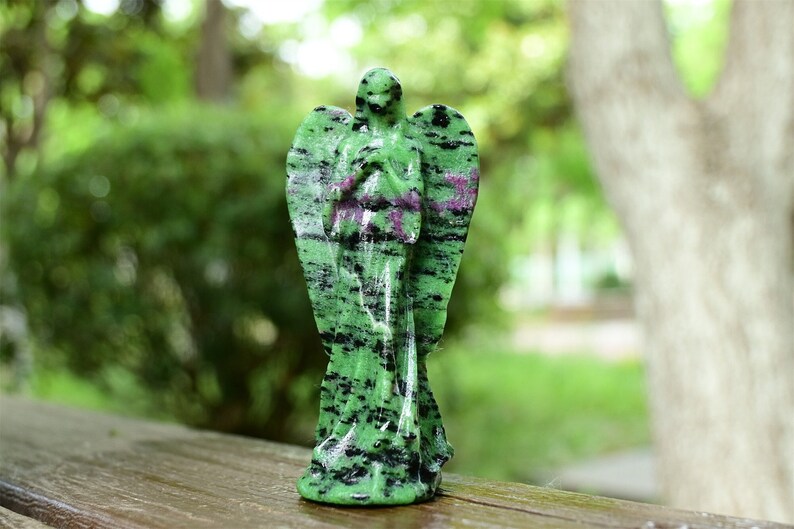 May include: Green and black gemstone angel figurine with a pink stripe. The angel is standing with its hands clasped in front of it.