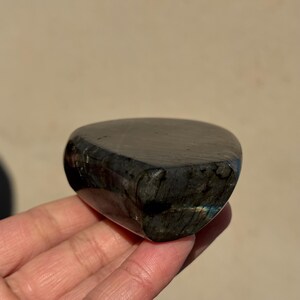 May include: A dark gray, polished labradorite stone with iridescent blue and green flashes. The stone is shaped like a heart and has a smooth, rounded surface.