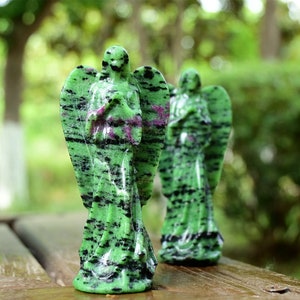 May include: Two green and black angel figurines carved from green aventurine gemstone. The figurines are standing on a wooden surface.