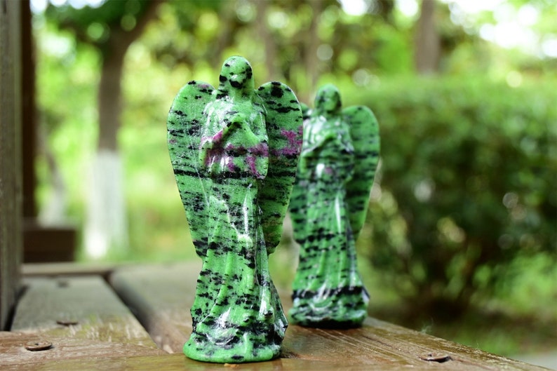 May include: Two green and black angel figurines carved from green aventurine gemstone. The figurines are standing on a wooden surface.
