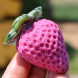 May include: A close-up of a pink strawberry with a green leafy top. The strawberry has a textured surface with small indentations. The green leaves have white and blue accents. The background is blurred.